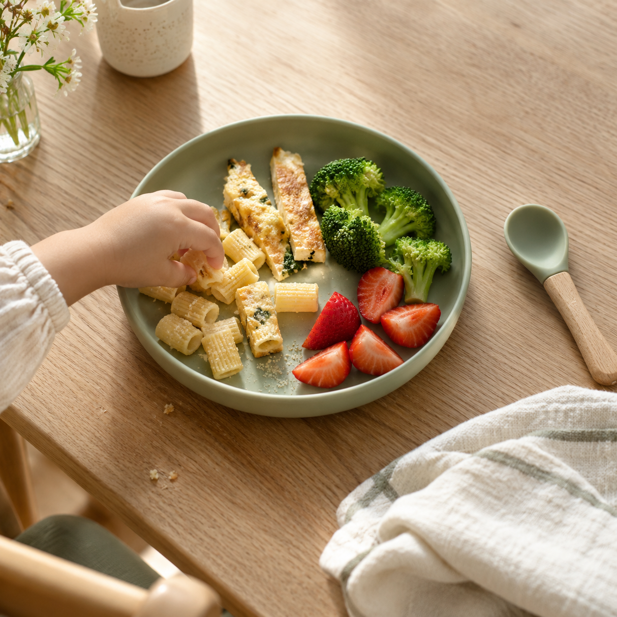A child's hand reaching for pasta on a green plate with vegetables, egg and strawberries